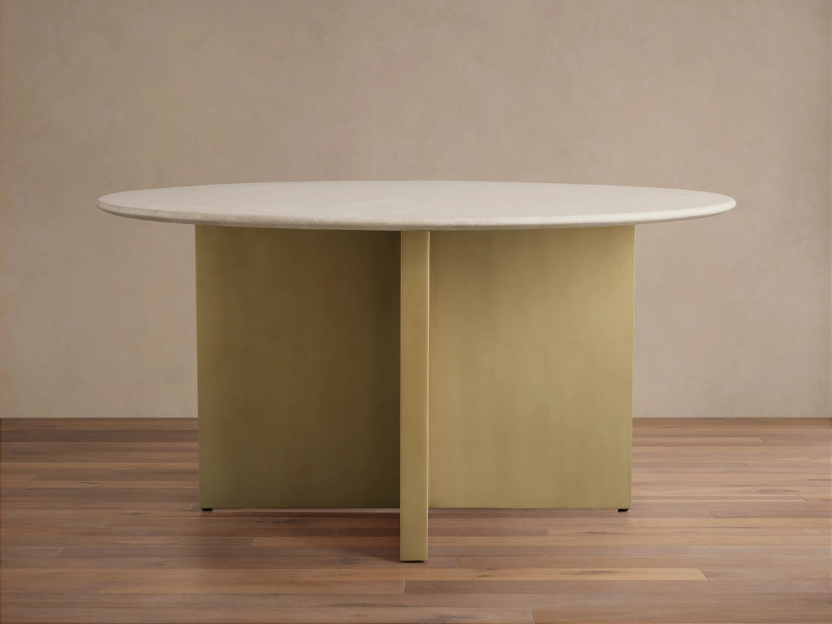 Saint Germain Round Stone Dining Table with light stone tabletop and brass-colored base, set on wooden floor against a beige wall.