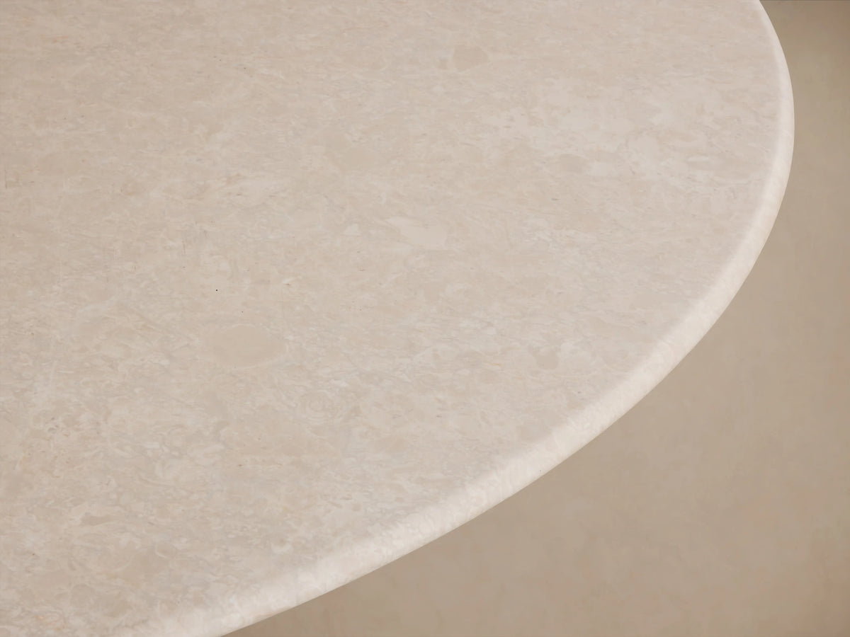 Round dining table with a cream marble top and a sleek, flared steel base in a dark finish, on a wood floor against a beige wall.