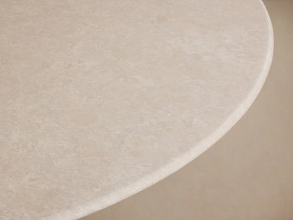 Round dining table with a cream marble top and a sleek, flared steel base in a dark finish, on a wood floor against a beige wall.