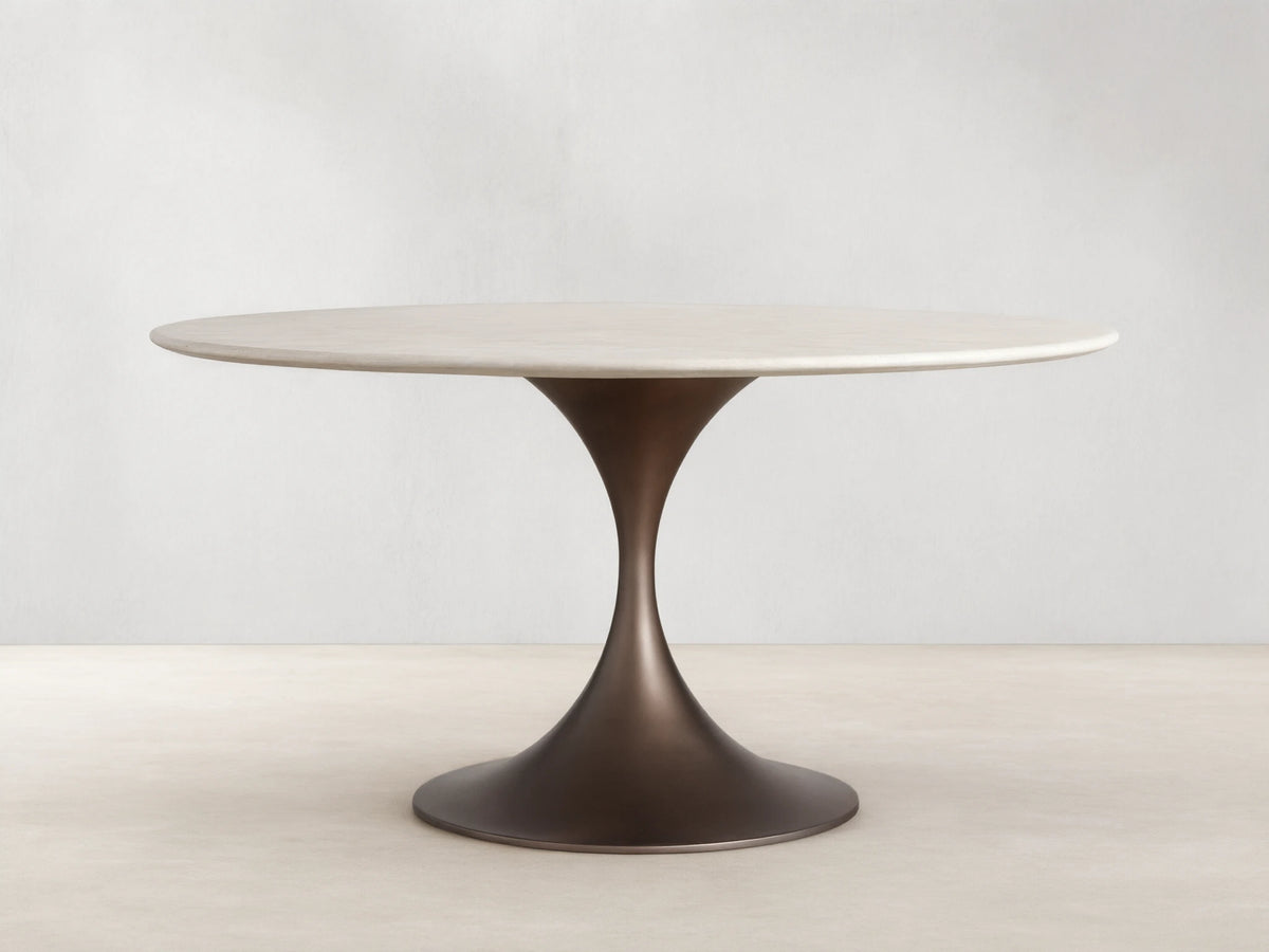 Round dining table with a cream marble top and a sleek, flared steel base in a dark finish, on a wood floor against a beige wall.