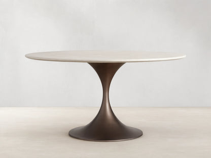 Round dining table with a cream marble top and a sleek, flared steel base in a dark finish, on a wood floor against a beige wall.