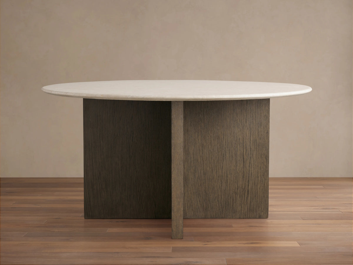 Round dining table with a smooth Carrara marble top. The wood base is a cross design in a chestnut finish, set on a wooden floor.