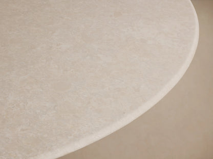 Round dining table with a smooth beige stone top and a golden rectangular base. The table sits on a wooden floor against a neutral wall.