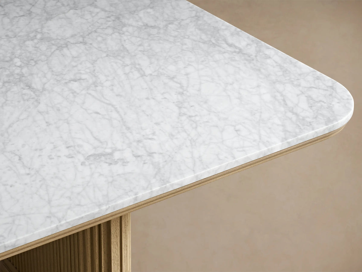 Close-up of the Coventry Dining Table's corner. Honed white Carrara marble top with gray veining, rounded edge, and light oak reeded base.