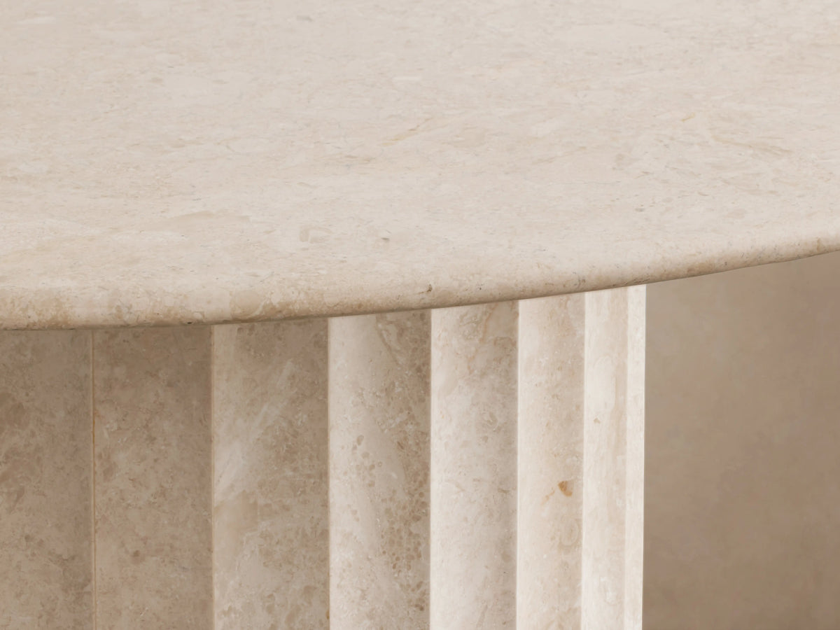 Danae Round Stone Dining Table. Smooth, light beige round marble top on a ribbed cylindrical stone base, on wood floor.