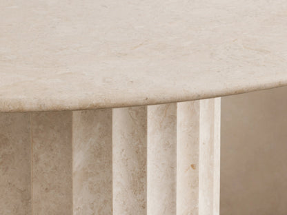 Danae Round Stone Dining Table. Smooth, light beige round marble top on a ribbed cylindrical stone base, on wood floor.