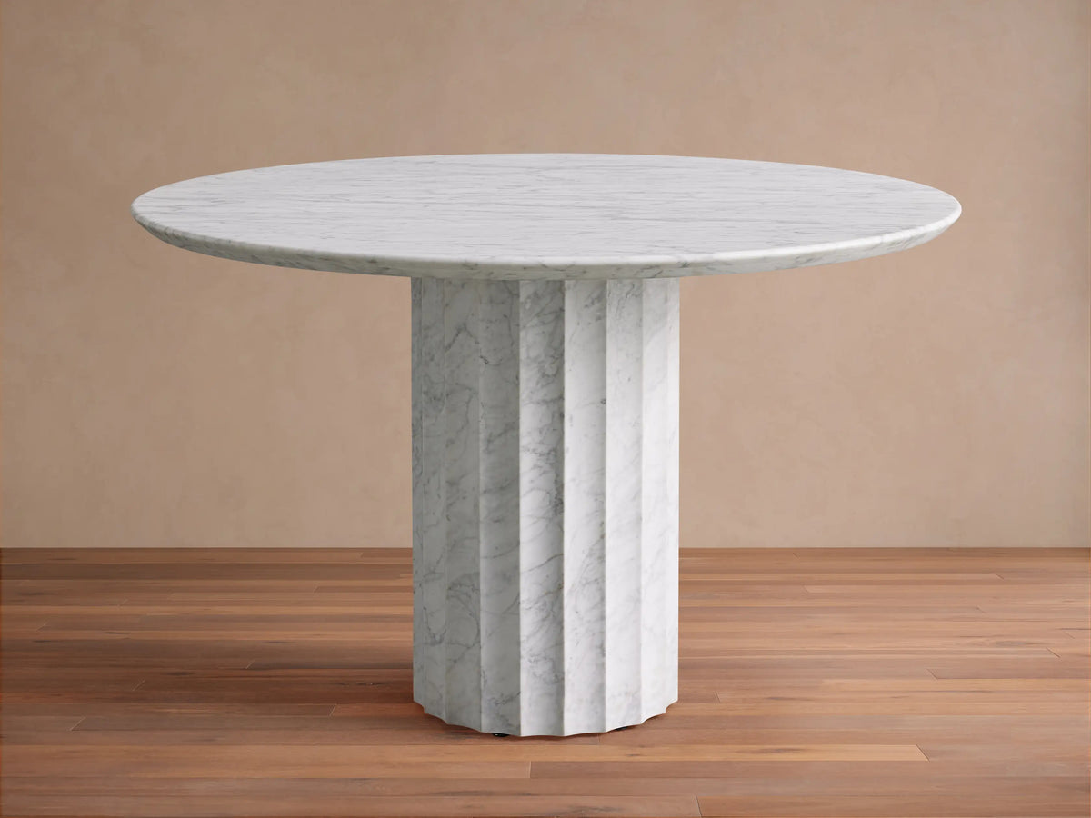 The Danae Round Stone Dining Table is shown, featuring a white marble top and a fluted pedestal base, set on a wooden floor.