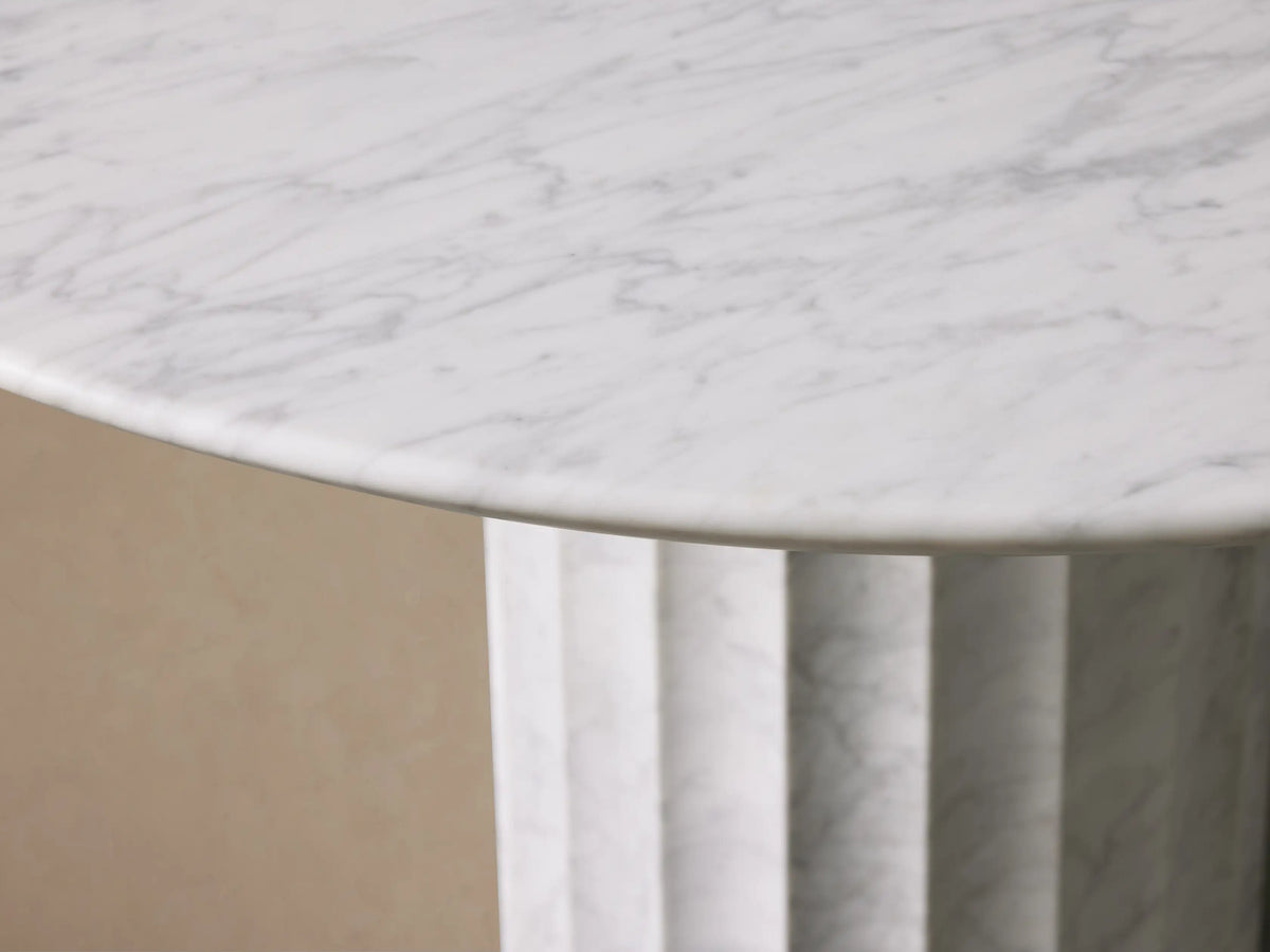 The Danae Round Stone Dining Table is shown, featuring a white marble top and a fluted pedestal base, set on a wooden floor.