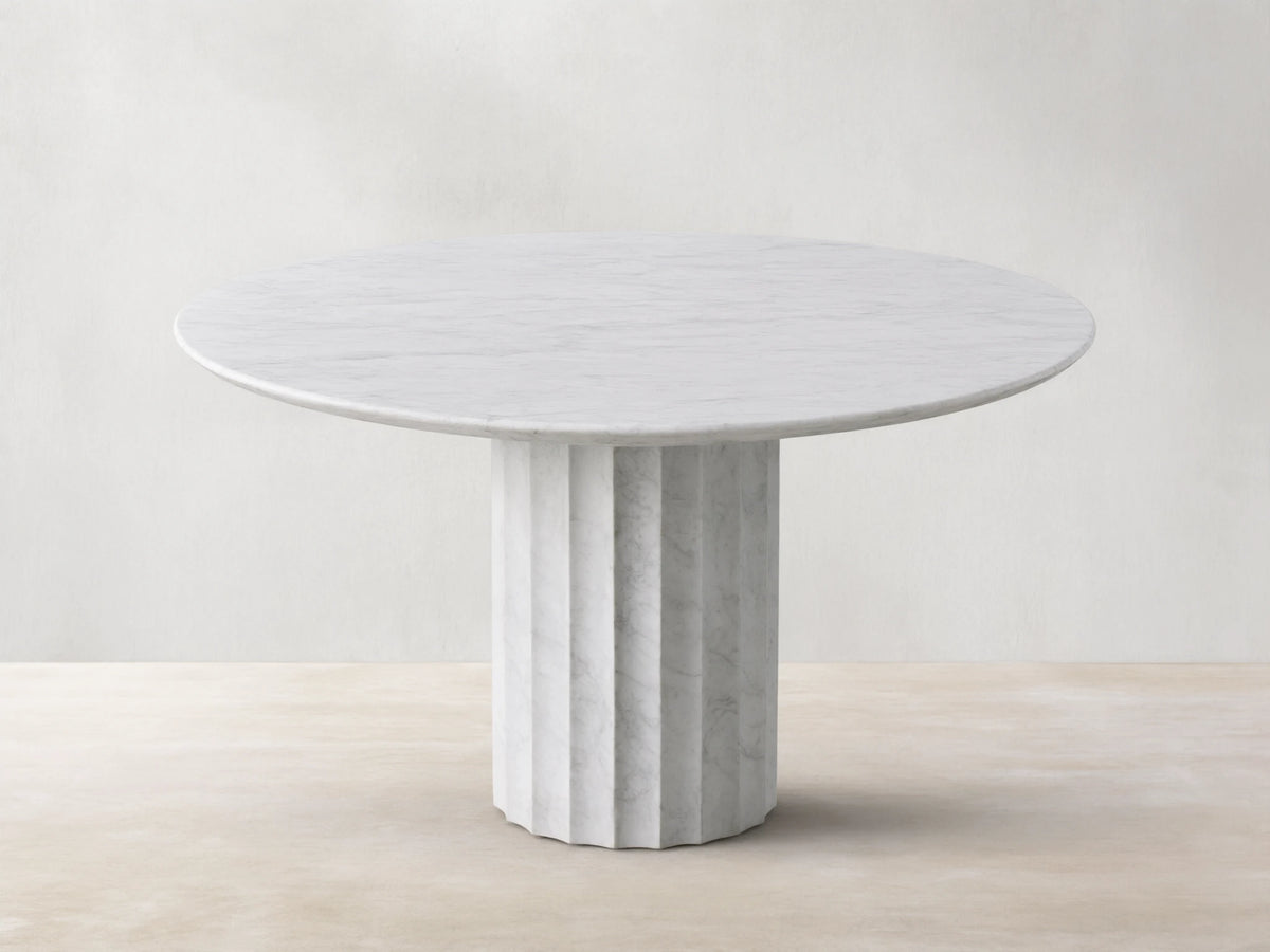 The Danae Round Stone Dining Table is shown, featuring a white marble top and a fluted pedestal base, set on a wooden floor.