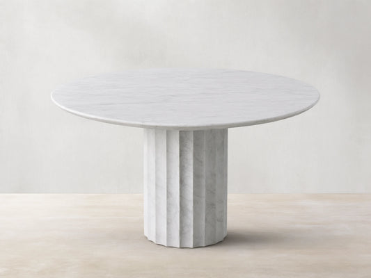 The Danae Round Stone Dining Table is shown, featuring a white marble top and a fluted pedestal base, set on a wooden floor.