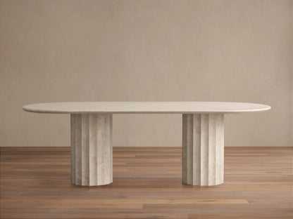 Danae Stone Dining Table in grey marble with oval top and dual fluted pedestal bases, set on wooden floor against beige wall.