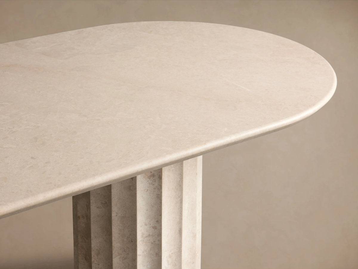 Danae Stone Dining Table with a smooth marble surface and two fluted column bases, set on a wooden floor against a beige wall.