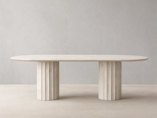 Danae Stone Dining Table in grey marble with oval top and dual fluted pedestal bases, set on wooden floor against beige wall.