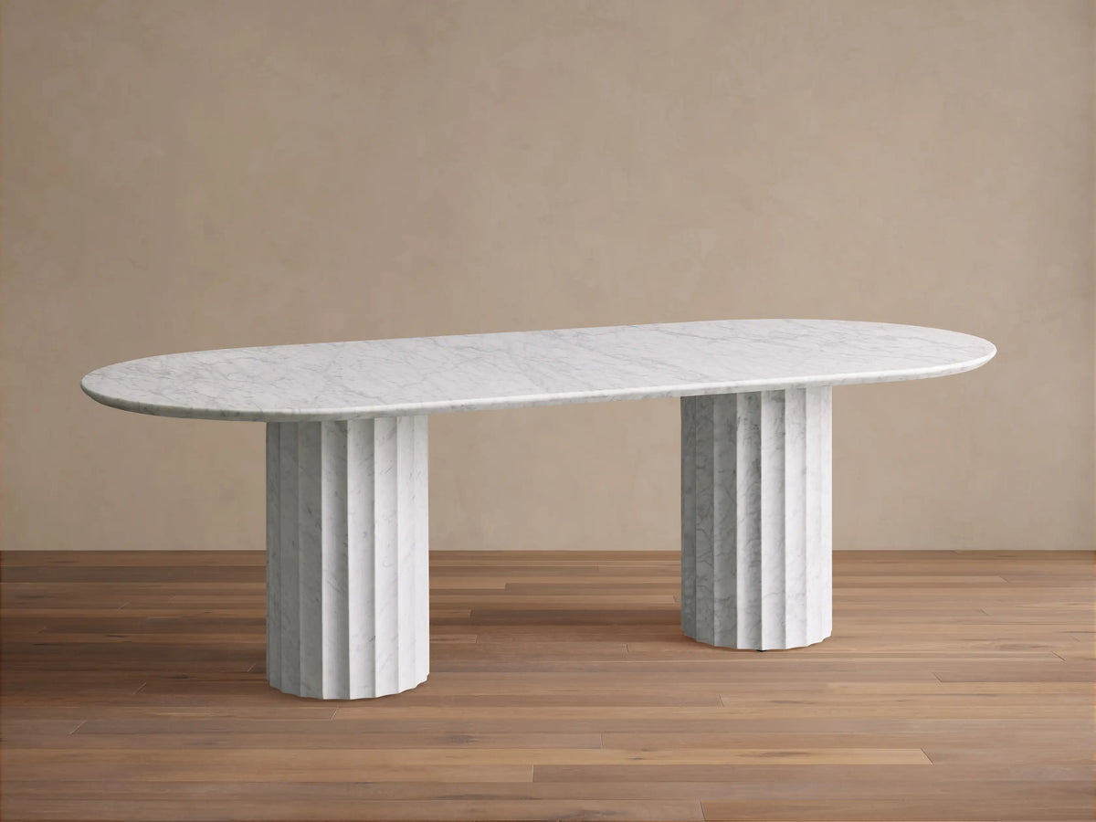 Danae Stone Dining Table: White oval marble top with subtle grey veining, resting on two fluted marble bases on a wood floor.
