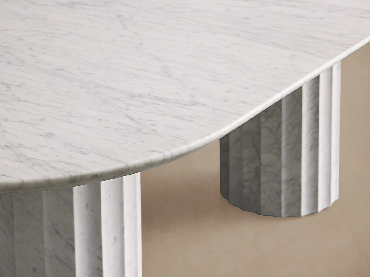 Danae Stone Dining Table, oval white marble top on two fluted marble cylindrical bases. Light tan wall and wood floor background.