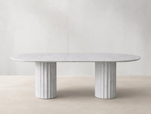 Danae Stone Dining Table: White oval marble top with subtle grey veining, resting on two fluted marble bases on a wood floor.