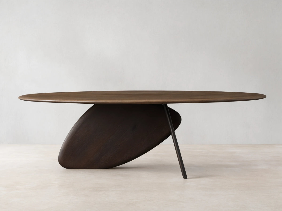 Pebble Dining Table with an oval russet oak top and a dark espresso sculptural, pebble-shaped base with a metal leg.