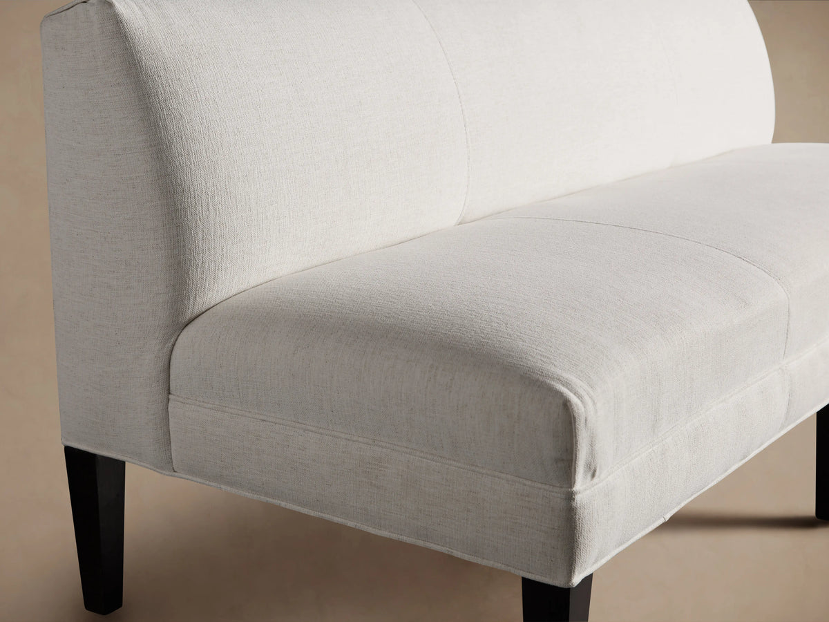 Eaton Settee in soft, pale fabric with a sleek, armless design. Features three sections and dark, tapered maple legs on a wooden floor.