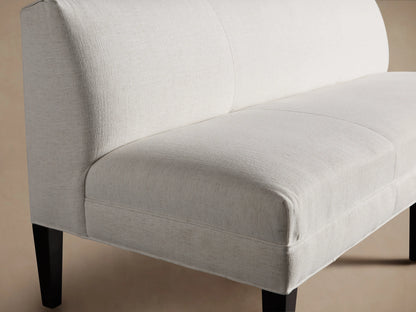 Eaton Settee in soft, pale fabric with a sleek, armless design. Features three sections and dark, tapered maple legs on a wooden floor.