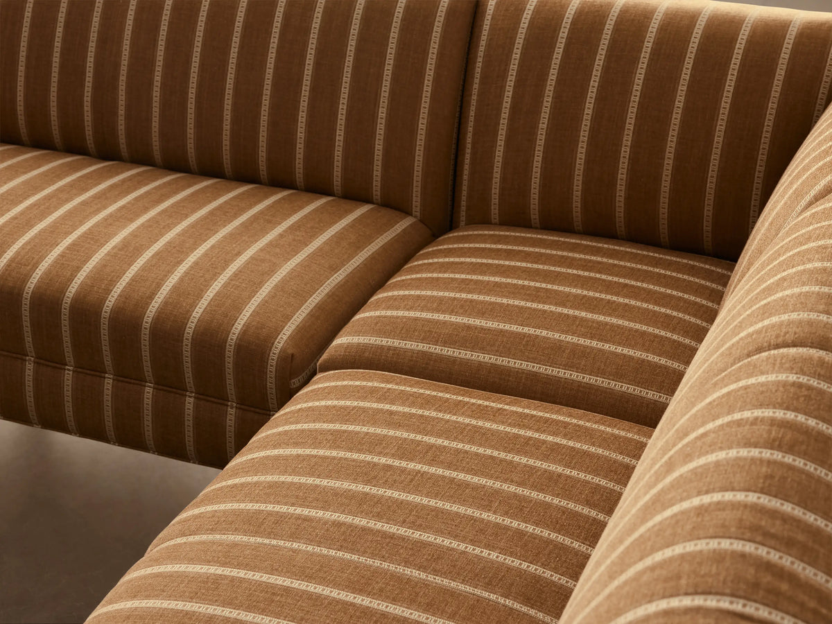 L-shaped Eaton Banquette in golden-brown striped fabric with dark tapered legs, on a light wood floor against a plain wall.