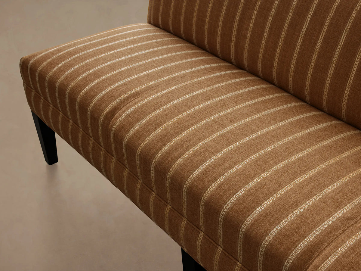 Eaton Settee, golden-brown fabric with light vertical stripes, dark wooden legs, on a wood floor against a light wall.