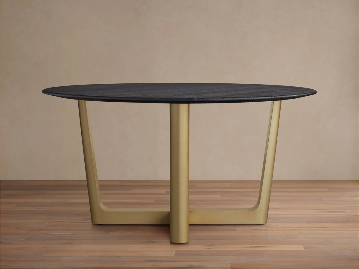 The Saint Germain Round Wood Dining Table features a dark oak tabletop and a wide, V-shaped Rennes base in antique brass on a wood floor.