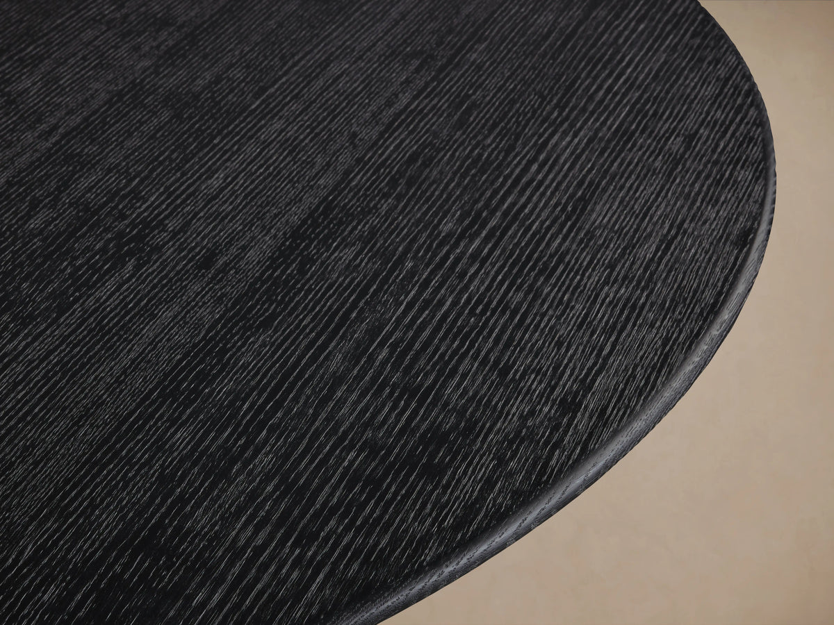 Round dining table with dark oak top and a U-shaped bronze base on a wooden floor. Neutral wall in the background.