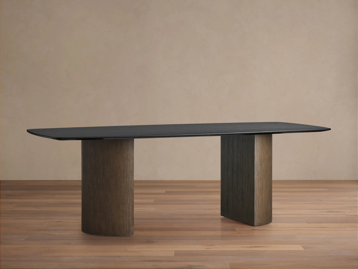 Saint Germain Wood Dining Table with an Ebony finish, features a sleek black top and two cylindrical wooden bases on a wood floor.