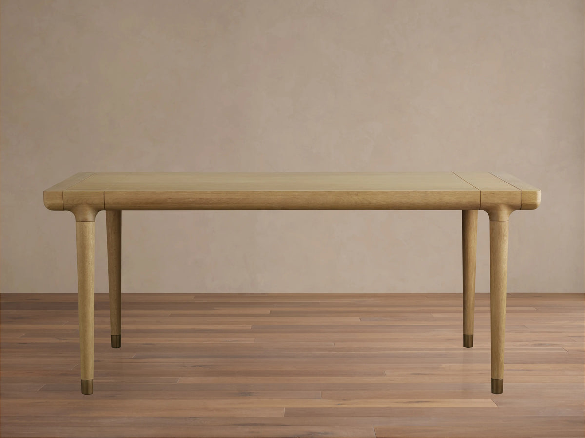 Eletta Dining Table, light oak, rectangular with rounded corners. Tapered legs with brass-colored caps, set on a wood floor.