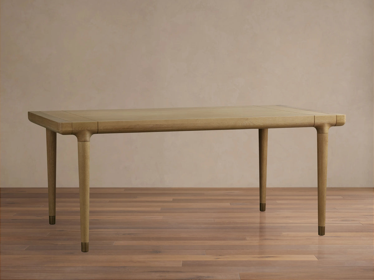 Eletta Dining Table, light oak, rectangular with rounded corners. Tapered legs with brass-colored caps, set on a wood floor.