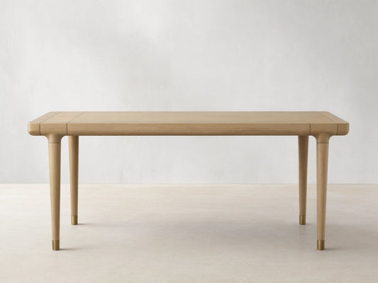 Eletta Dining Table, light oak, rectangular with rounded corners. Tapered legs with brass-colored caps, set on a wood floor.