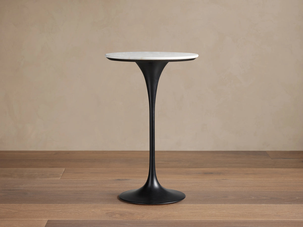 Enzo Bar Table with a round, polished white marble tabletop on a sleek, black tulip-style iron base, set against a wooden floor.