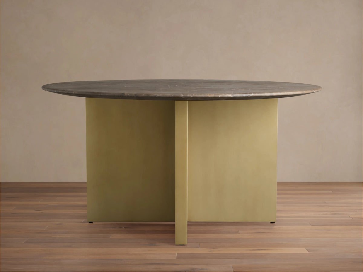 Saint Germain Round Stone Dining Table with a dark stone top and gold-colored V-shaped base on a wooden floor.