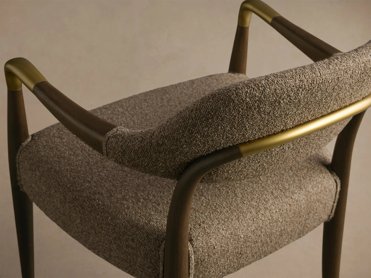 Close-up, angled view of the Evelyn Dining Arm Chair, with dark mahogany wood frame, brushed brass accents, and textured brown upholstery.