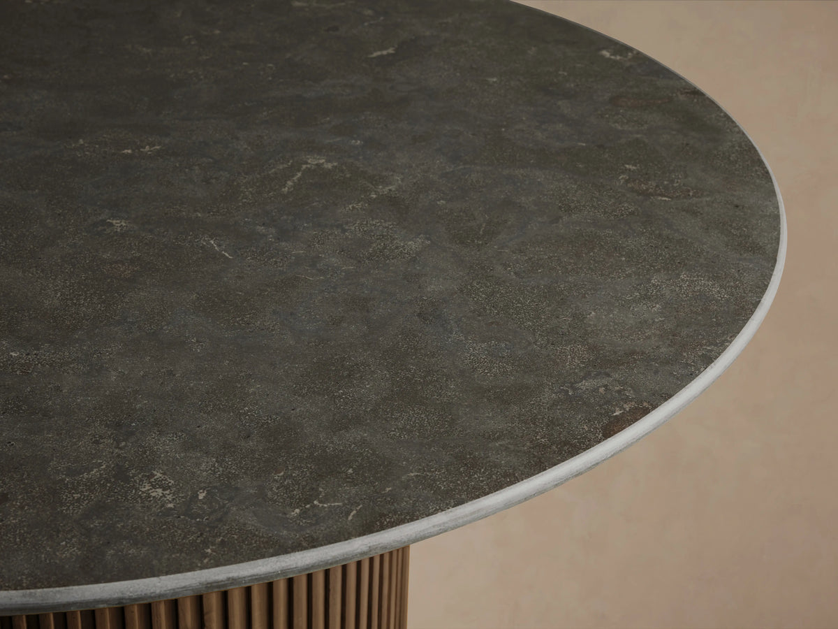 Finnley Round Dining Table with a smooth dark marble top and ribbed cylindrical base in a brass finish, on a wooden floor.