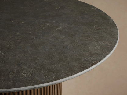 Finnley Round Dining Table with a smooth dark marble top and ribbed cylindrical base in a brass finish, on a wooden floor.