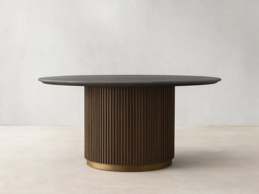 Finnley Round Dining Table with a smooth dark marble top and ribbed cylindrical base in a brass finish, on a wooden floor.
