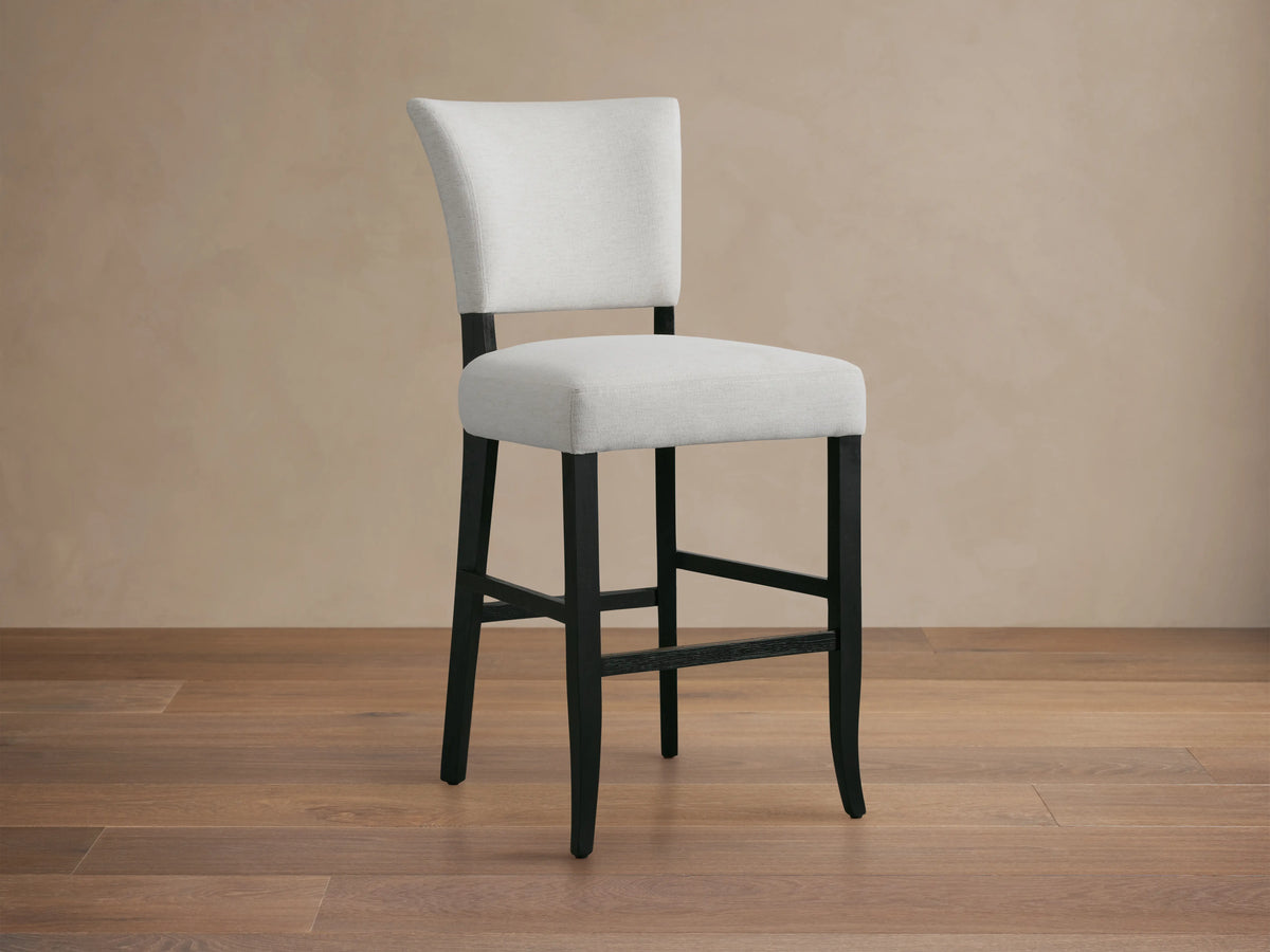 Fallyn Stool with an upholstered, light fabric seat and backrest, crafted from dark, painted wood. Positioned on wooden flooring.