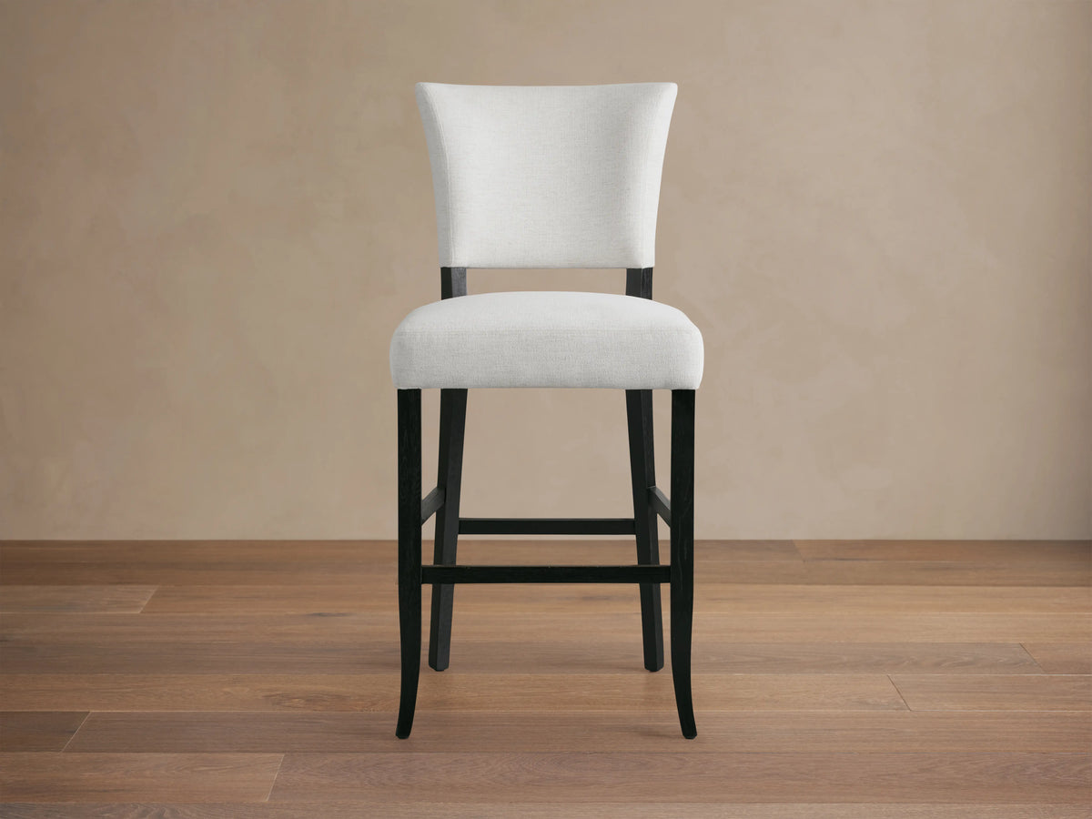 Fallyn Stool with white cushioned seat and backrest, black wooden legs on hardwood floor, against a beige wall.