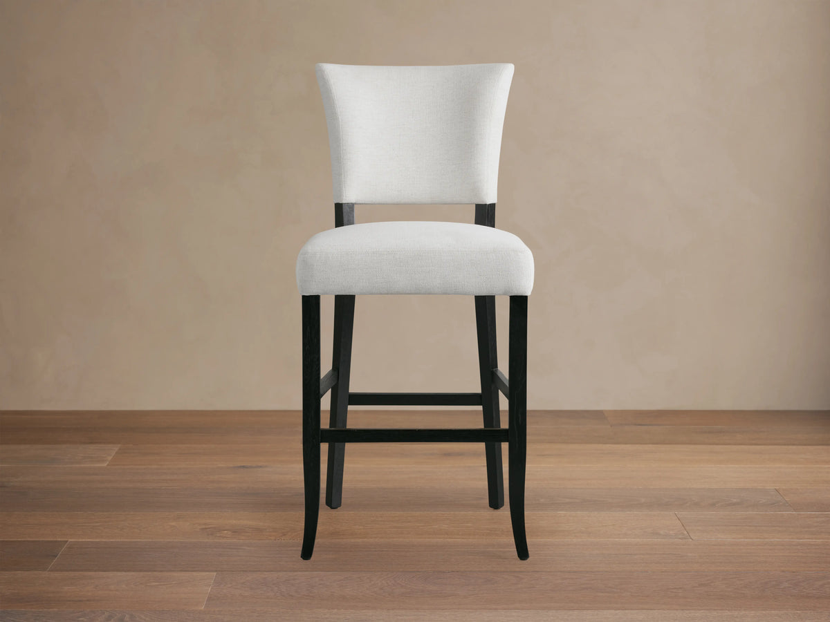 Fallyn Stool with an upholstered, light fabric seat and backrest, crafted from dark, painted wood. Positioned on wooden flooring.