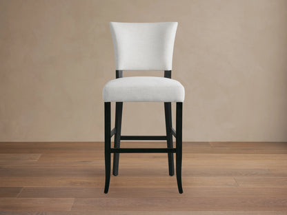 Fallyn Stool with an upholstered, light fabric seat and backrest, crafted from dark, painted wood. Positioned on wooden flooring.
