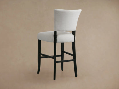 A close-up of the Fallyn Stool showing the soft white fabric seat and backrest, supported by black solid wood legs. The texture of the fabric and the sturdy frame are visible.