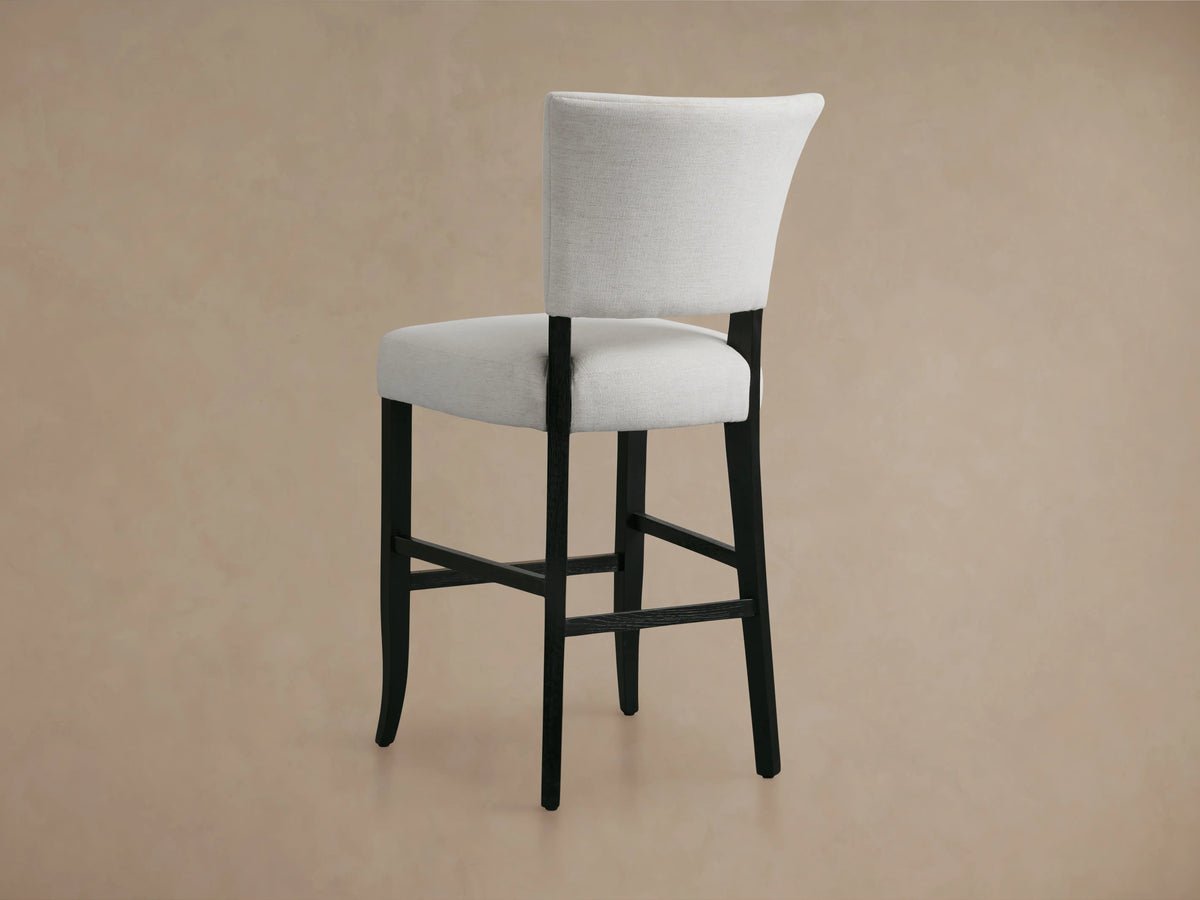 The Fallyn Stool features a light gray upholstered back and seat with dark wooden legs, set against a neutral background.