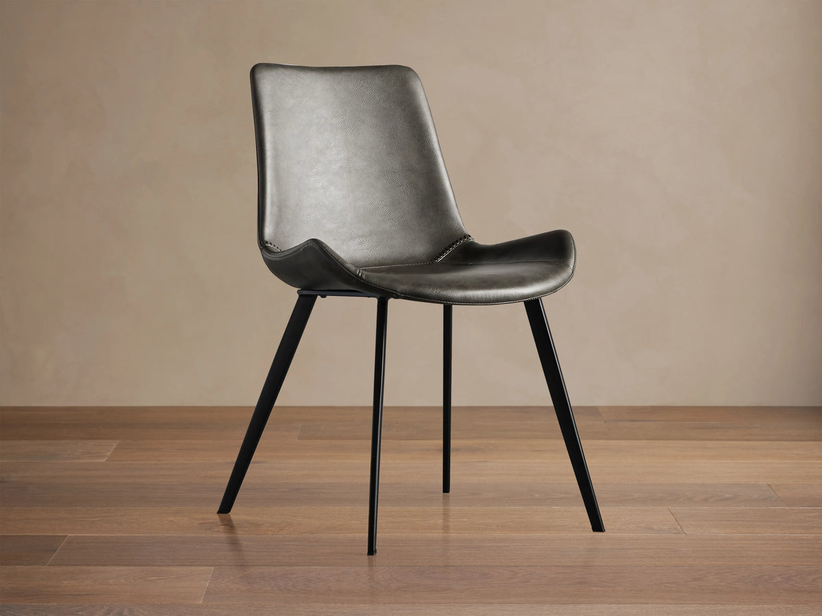 Angled Gage Faux Leather Dining Chair with dark gray seat, baseball stitching, and black iron legs on a wooden floor.