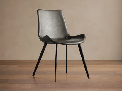 Angled Gage Faux Leather Dining Chair with dark gray seat, baseball stitching, and black iron legs on a wooden floor.