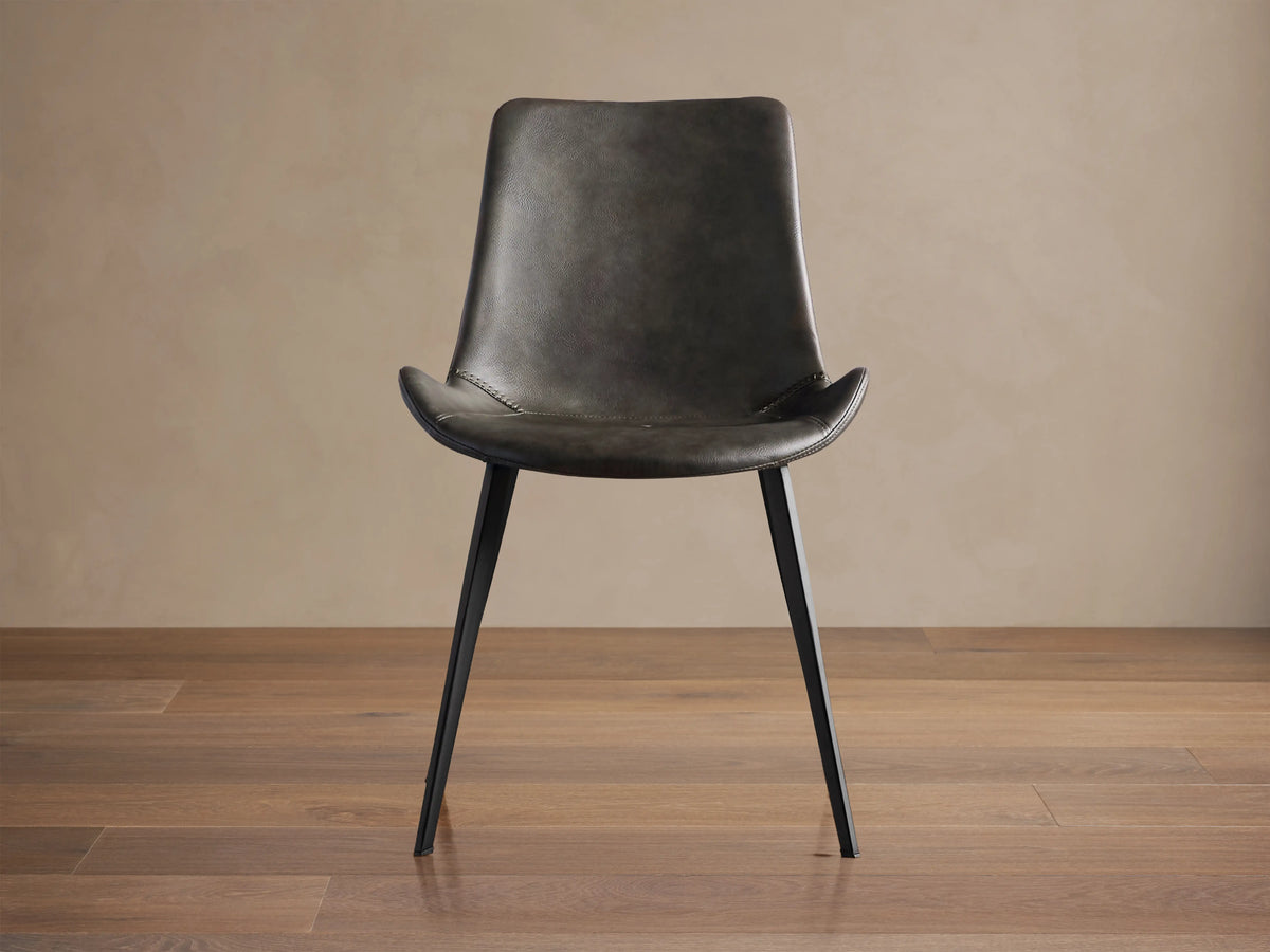 Angled Gage Faux Leather Dining Chair with dark gray seat, baseball stitching, and black iron legs on a wooden floor.