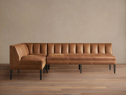Gates L-shaped leather banquette in distressed caramel brown with channel-stitched backs, dark tapered legs, on wood floor.