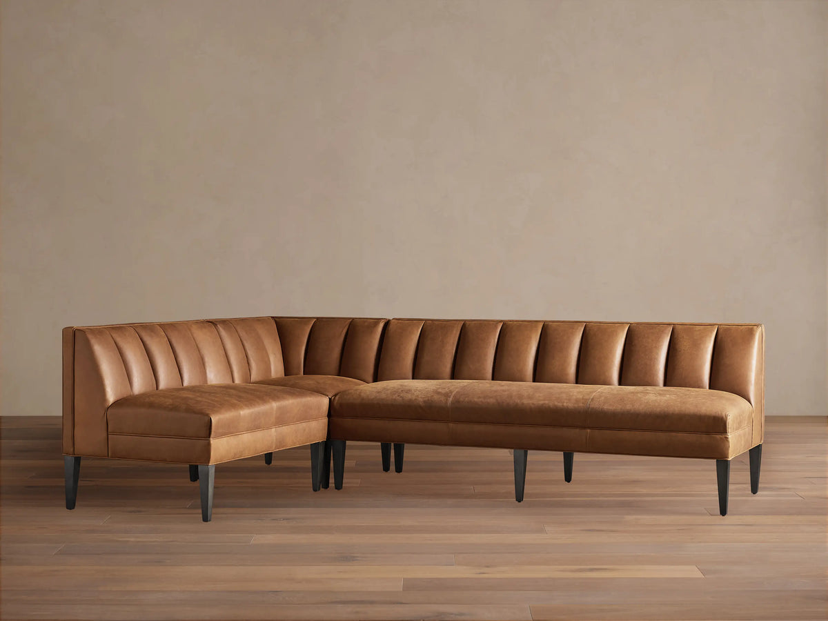 Gates L-shaped leather banquette in distressed caramel brown with channel-stitched backs, dark tapered legs, on wood floor.