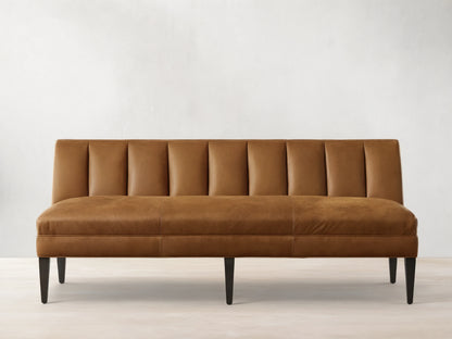 Gates Leather Settee, a tan leather bench with a channel-tufted backrest, smooth seat, and dark tapered legs, against a light wall.