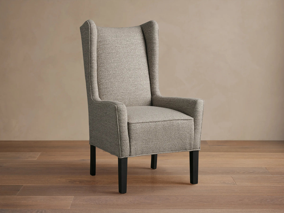 Halstead Dining Chair in gray fabric featuring a high wingback design, padded seat, and black wooden legs, set on a wooden floor.